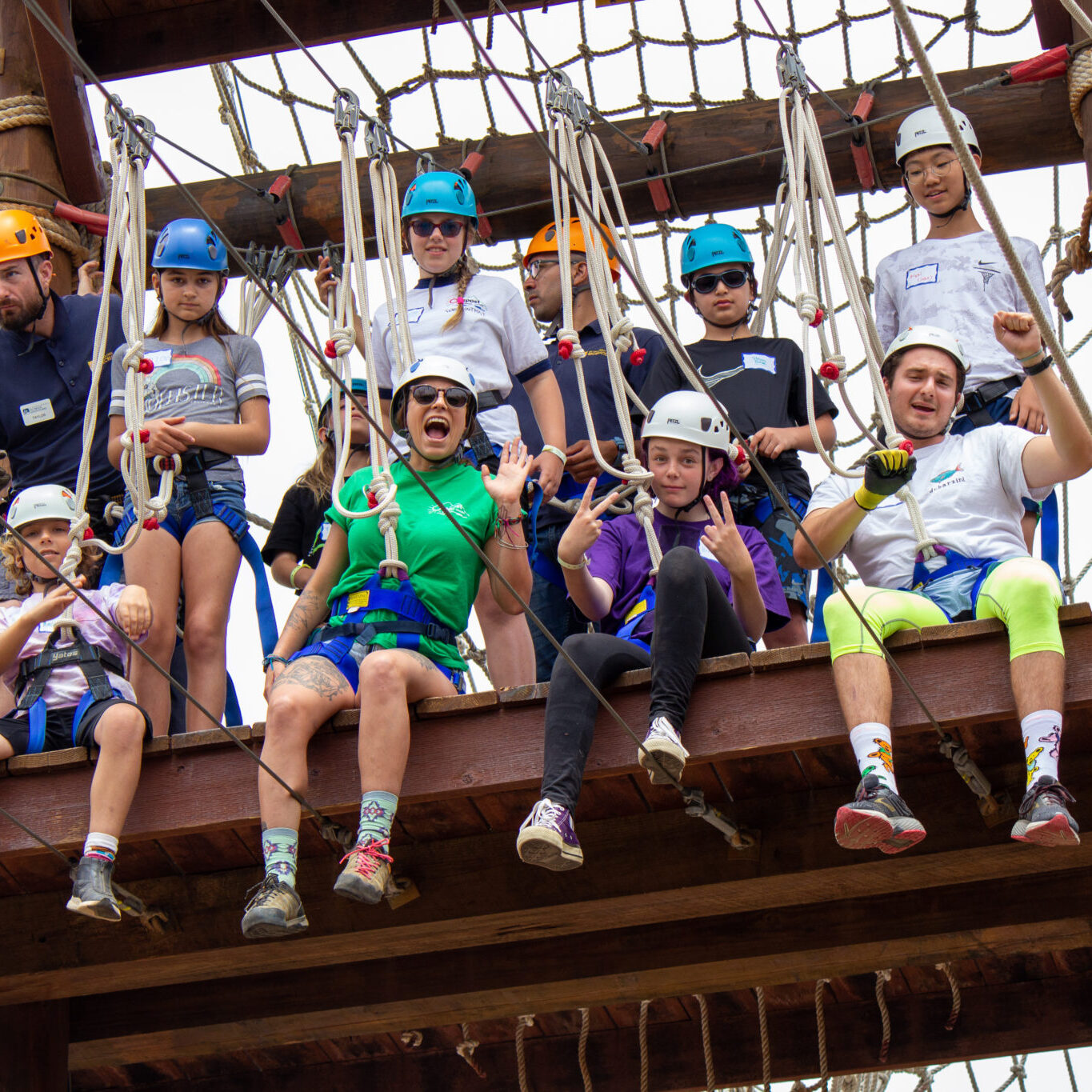 Campers and instructors wearing helmets and safety harnesses prepare on a wooden ropes-course structure.